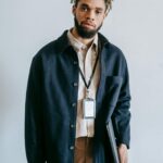 Confident young African American male entrepreneur with stylish haircut and beard in casual clothes and name tag standing against white wall with laptop and bag in hands