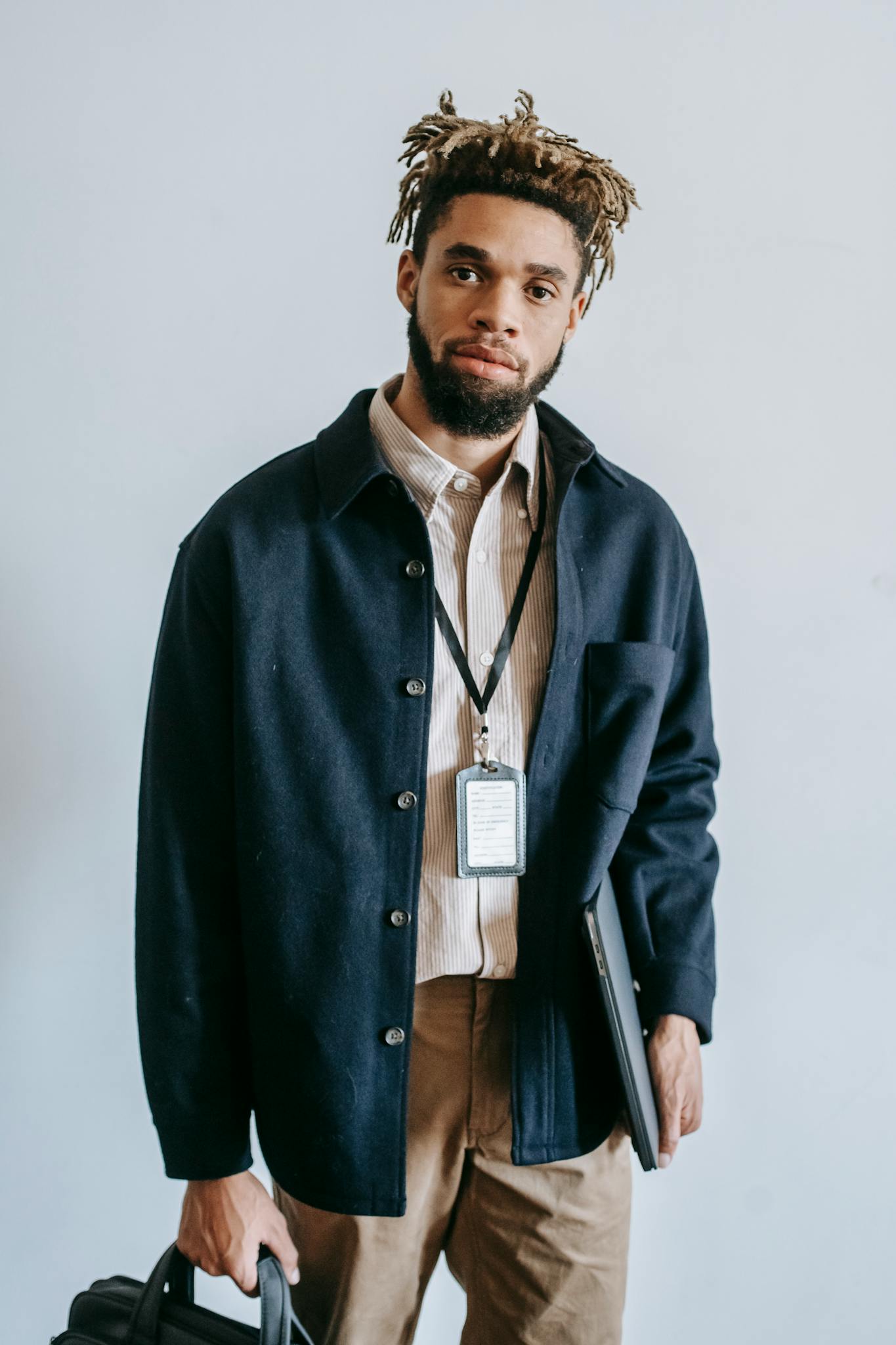 Confident young African American male entrepreneur with stylish haircut and beard in casual clothes and name tag standing against white wall with laptop and bag in hands